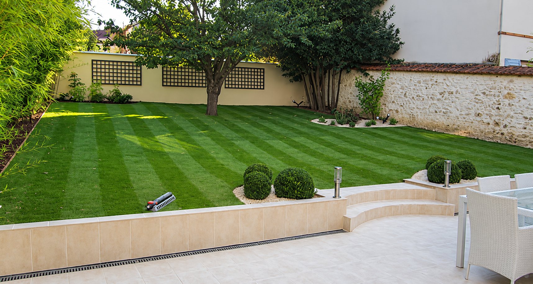 Un jardin clos avec une pelouse tondue en bandes, des buis taillés en boule, des marches arrondies et une terrasse carrelée meublée de fauteuils blancs.