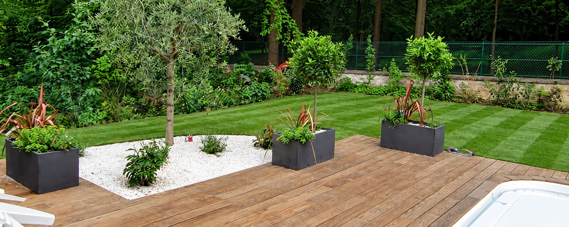 Jardin récemment aménagé avec une terrasse en bois, des jardinières grises, un olivier dans un massif de gravier blanc et une pelouse fraîchement tondue.