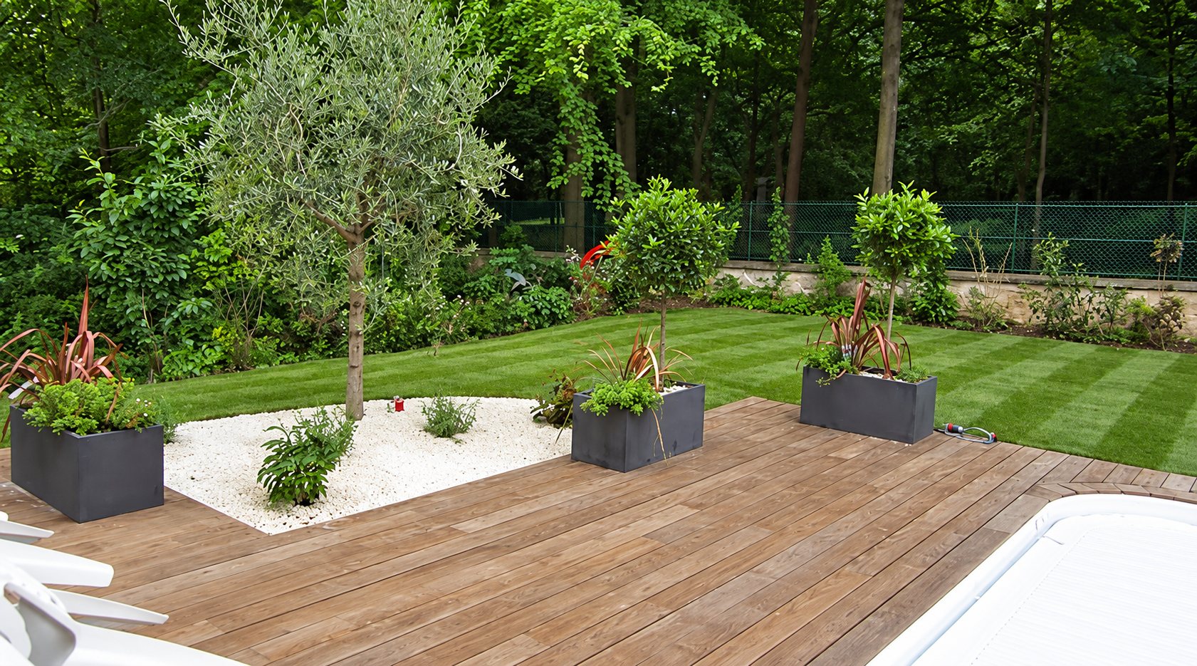 Une terrasse en bois exotique entourée d'un jardin aménagé avec un olivier, des bacs gris contenant des plantes, un carré de galets blancs et une pelouse tondue en bandes.